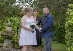 smiling bride & groom holding hands in garden with celebrant looking on